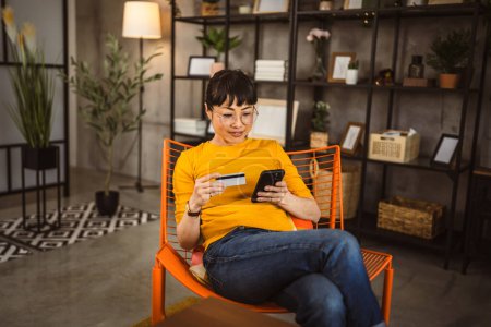 Foto de Japonesa madura mujer con gafas se sientan en silla de uso de tarjeta de crédito y teléfono móvil en línea de compras en casa - Imagen libre de derechos