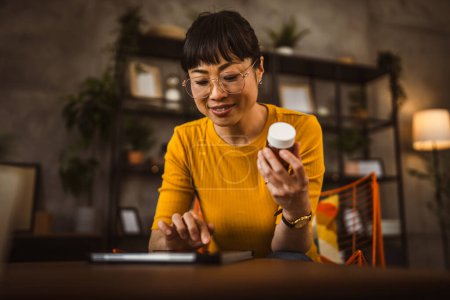 Téléchargez les photos : Japonaise mature femme avec des lunettes de vue assis sur la chaise et lire la recette de médicament utilisation tablette à la maison - en image libre de droit