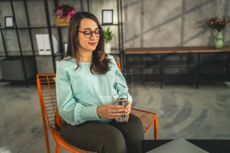 Photo for Adult caucasian pregnant women wear eyeglasses sit on chair and hold glass of water at home - Royalty Free Image