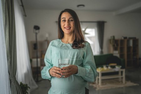 Photo for Adult caucasian pregnant women wear eyeglasses sit on chair and hold glass of water at home - Royalty Free Image