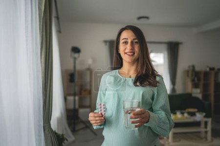 Téléchargez les photos : Les femmes enceintes caucasiennes adultes se tiennent debout et tiennent un médicament et un verre d'eau à la maison - en image libre de droit