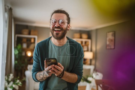 Téléchargez les photos : Un homme caucasien adulte avec des lunettes barbe, tatouages et cheveux écrire un message ou naviguer sur Internet à la maison heureux - en image libre de droit