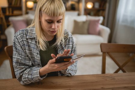 Téléchargez les photos : Jeune femme caucasienne utilisant le téléphone intelligent et les achats en ligne, tenant la carte de crédit effectuant le paiement assis au bureau à la maison - en image libre de droit