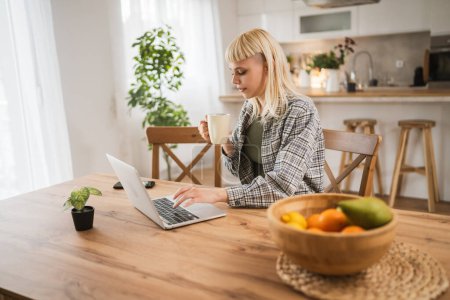 Foto de Joven mujer rubia caucásica trabajar en el ordenador portátil y sostener la taza de casa feliz positivo - Imagen libre de derechos