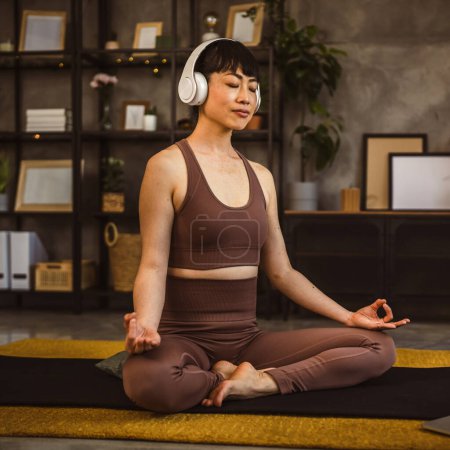 Eine Japanerin auf dem Boden zu Hause praktiziert Yoga online geführte Meditation mit Kopfhörern auf dem Kopf mit Laptop-Computer