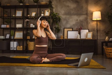 Foto de Una mujer japonesa en el suelo en casa practica yoga en línea meditación guiada con auriculares en su cabeza use computadora portátil - Imagen libre de derechos
