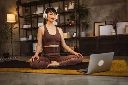 Téléchargez les photos : Une femme japonaise sur le sol à la maison pratique le yoga méditation guidée en ligne avec des écouteurs sur sa tête utiliser ordinateur portable - en image libre de droit