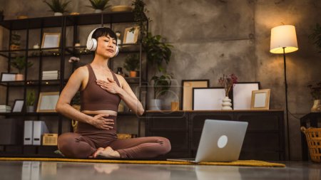 Eine Japanerin auf dem Boden zu Hause praktiziert Yoga online geführte Meditation mit Kopfhörern auf dem Kopf mit Laptop-Computer