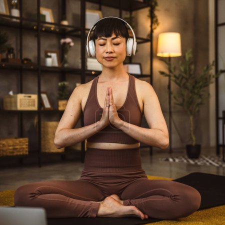Foto de Una mujer japonesa en el suelo en casa practica yoga en línea meditación guiada con auriculares en su cabeza use computadora portátil - Imagen libre de derechos