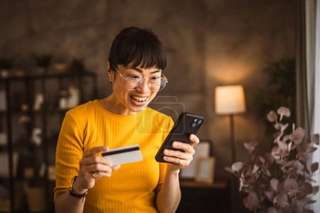 Téléchargez les photos : Japonais mature femme acheter en ligne avec téléphone mobile et carte de crédit à la maison - en image libre de droit