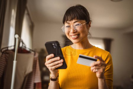 Téléchargez les photos : Japonais mature femme acheter en ligne avec téléphone mobile et carte de crédit à la maison - en image libre de droit