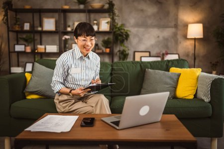 Téléchargez les photos : Femme japonaise mature tenir presse-papiers et regarder ordinateur portable pendant le travail de la maison - en image libre de droit