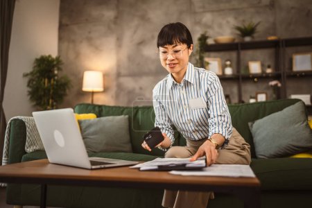 Téléchargez les photos : Femme japonaise mature avec des lunettes tenir téléphone mobile et regarder document travail de la maison - en image libre de droit