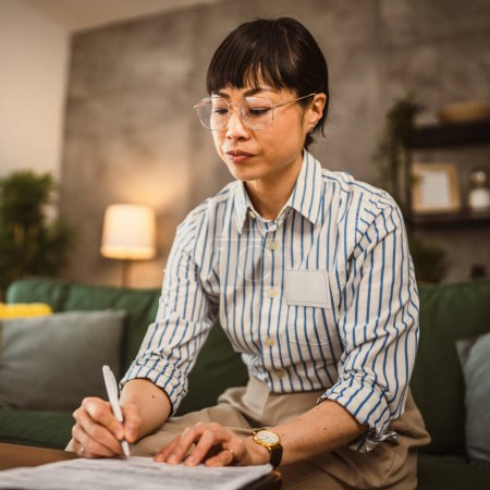 Téléchargez les photos : Femme japonaise mature avec des lunettes de travail de la maison et signer le document - en image libre de droit
