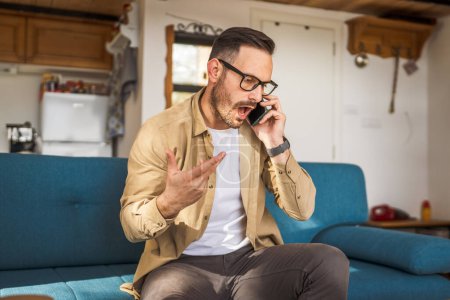 Hombre enojado adulto sentarse en el sofá y hablar en el teléfono móvil en el trabajo en casa desde casa