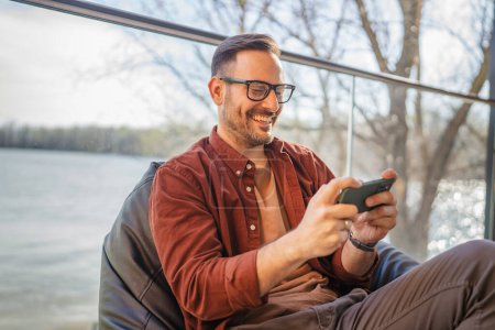 Foto de Adulto caucásico con gafas hombre jugar videojuegos en su teléfono en casa - Imagen libre de derechos