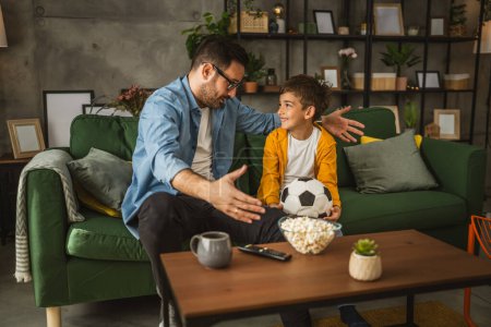 Téléchargez les photos : Père et fils caucasien regarder match de football et acclamer à la maison bruyant, excité, heureux - en image libre de droit