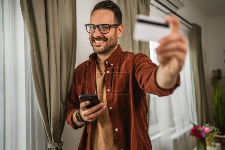 Téléchargez les photos : Portrait d'homme adulte boutique en ligne avec téléphone portable et détenir une carte de crédit à la maison - en image libre de droit