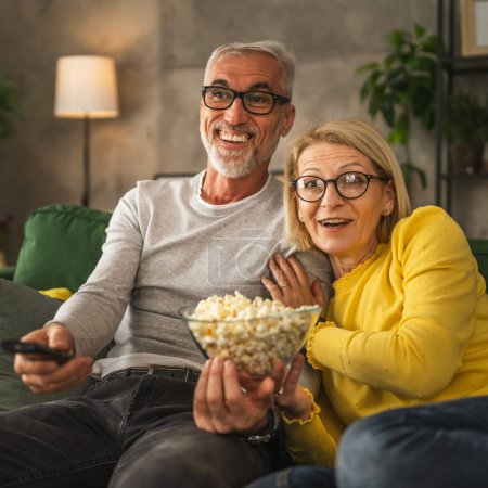 Photo for Mature senior couple man and woman husband and wife watch movie and eat popcorn with remote control at home - Royalty Free Image