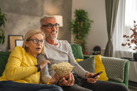Photo for Mature senior couple husband and wife watch scary movie and hold popcorn with remote control at home - Royalty Free Image