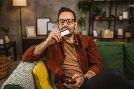 Téléchargez les photos : Portrait de l'homme adulte baiser carte de crédit après avoir réussi les achats en ligne à la maison - en image libre de droit