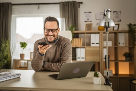 Foto de Hombre adulto trabaja en el ordenador portátil y grabar correo de voz o mensaje en el teléfono celular en la oficina - Imagen libre de derechos