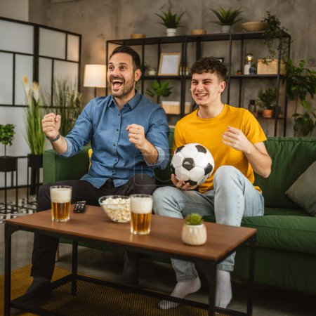 Téléchargez les photos : Jeune caucasien et adulte caucasien homme frères ou amis regarder le football et le match de cheer tenir ballon de football avec du maïs soufflé et de la bière sur la table à la maison - en image libre de droit