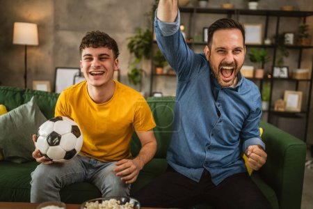 Téléchargez les photos : Young caucasian and adult caucasian man brothers or friends watch football and cheer match hold football ball with popcorn and beer on table at home - en image libre de droit