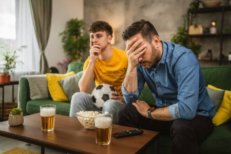 Téléchargez les photos : Deux hommes amis caucasiens sont déçus tout en regardant le match de football parce qu'ils équipe perdent le match à la maison - en image libre de droit