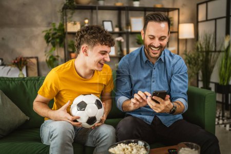 Zwei männliche Freunde verfolgen Ergebnisse von Fußballspielen zu Hause auf dem Handy