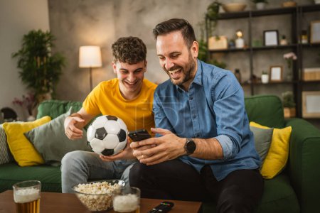 Two male friends follow football match results on mobile phone at home