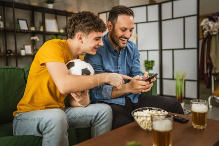 Zwei männliche Freunde verfolgen Ergebnisse von Fußballspielen zu Hause auf dem Handy