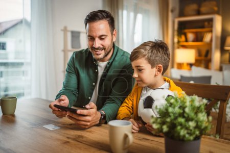 Téléchargez les photos : Père et fils caucasien regarder match de football sur téléphone mobile et acclamer à la cuisine bruyant, excité, heureux - en image libre de droit