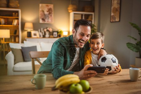Téléchargez les photos : Père et fils caucasien regarder match de football sur téléphone mobile et acclamer à la cuisine bruyant, excité, heureux - en image libre de droit