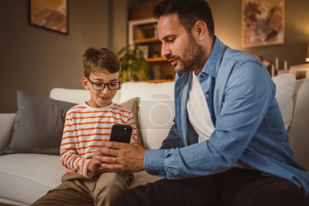 Father and son sit on sofa and use mobile phone at home have fun