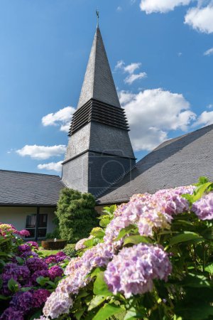 Téléchargez les photos : ODENTHAL, GERMANY - JULY 20, 2020: Parish church Saint Michael with blooming hortensia shrubs against blue sky on July 20, 2020 in Odenthal, Germany - en image libre de droit