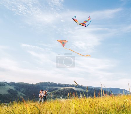 Smiling sister with launching brother with colorful kites - popular outdoor toy on the high grass hills meadow. Happy childhood moments or outdoor time spending concept image. 