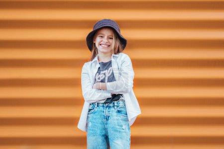 Photo for Little fashion-dressed girl portrait sincerely smiling while looking at camera on orange wall background. Urban people living and a street everyday life concept image - Royalty Free Image