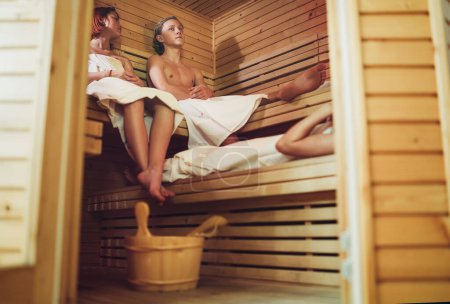 Foto de Relajante pareja de adolescentes envolvió sábanas blancas tumbadas y sudando en un banco de madera en la sauna finlandesa caliente disfrutando de un agradable tratamiento de temperatura corporal. Concepto de estilo de vida saludable. - Imagen libre de derechos