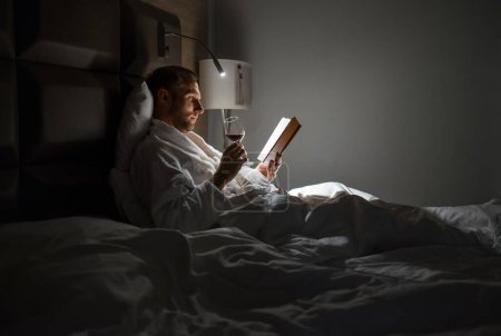 Photo for Midle-aged man relaxing rin bed reading book holding a glass of red wine with bedside lamp turned on. Evening relaxation, hobbies, free time concept. Adulthood concept. - Royalty Free Image