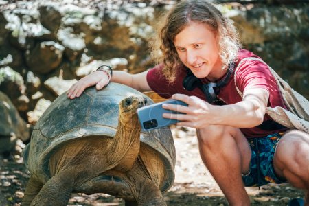 Photo for Smiling tourist boy making a selfie using cell phone with Aldabra giant tortoise endemic species - one of the largest tortoises in the world in the zoo nature park on Mauritius island. - Royalty Free Image