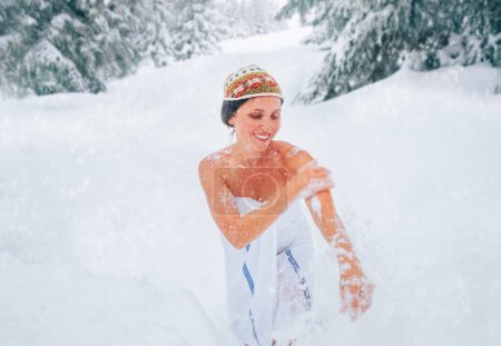 Mujer sonriente de mediana edad haciendo tratamiento de frotamiento de nieve corporal en la deriva profunda de nieve después de terminar sauna caliente. Cuerpo saludable endurecimiento templado y vacaciones de invierno en la imagen concepto de casa de campo.