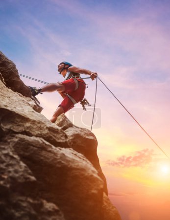 Hombre escalador musculoso en casco protector rappel de la pared de roca del acantilado usando cuerda dispositivo Belay y arnés de escalada en el fondo del cielo al atardecer. Concepto activo de gasto de tiempo en deportes extremos.
