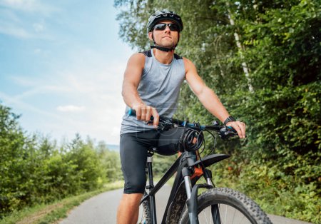 Photo for Portrait of happy man dressed in cycling clothes, helmet and sunglasses riding a bicycle on the out-of-town bicycle path. Active sporty people concept image. - Royalty Free Image