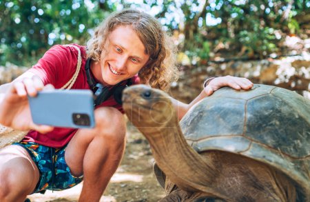 Smiling tourist boy making a selfie using cell phone with Aldabra giant tortoise endemic species - one of the largest tortoises in the world in the zoo nature park on Mauritius island.