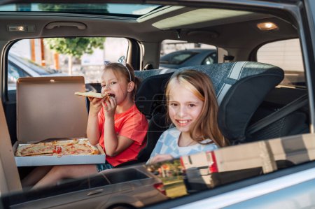 Photo for Two positive smiling sisters are happy to eat just cooked Italian pizza sitting in child car seats on the car back seat. Happy childhood, fast food eating, or auto journey lunch break concept image. - Royalty Free Image