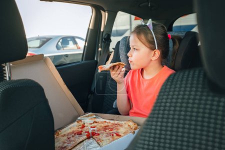 Photo for Portrait of little 5 YO girl eating just cooked Italian pizza sitting in child car seat on car back seat and looking out window . Happy childhood, fast food eating, or auto journey lunch break image - Royalty Free Image
