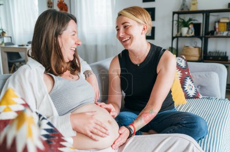Photo for Young woman tender touching partner's female pregnant belly. Same-sex marriage couple on home living room sofa.  Woman's health, happy pregnancy doula supporting and calm mental mood concept image - Royalty Free Image