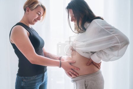 Young woman tender touching partner's female pregnant belly. Same-sex marriage couple next to living room window. Woman's health, happy pregnancy doula supporting and calm mental mood concept image.