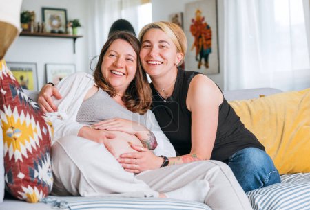 Photo for Happy smiling women couple waiting for baby. Same-sex pregnant marriage couple on home living room sofa.  Woman's health, happy pregnancy doula supporting and calm mental mood concept image - Royalty Free Image